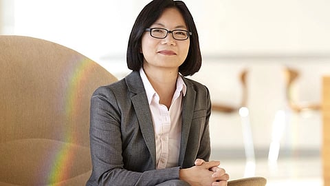A confident woman in a gray suit sits on a beige chair, with folded hands.