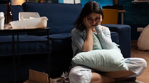 A woman sits on the floor looking despondent, hugging a pillow.