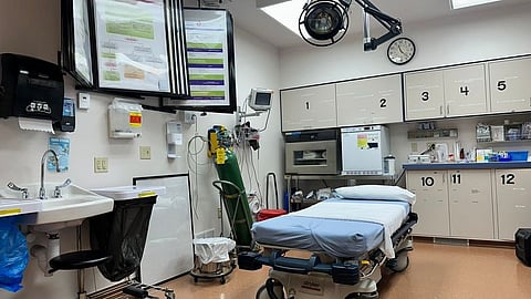 Hospital room featuring an empty medical bed, overhead light, wall-mounted monitors, and cabinets labeled with numbers.
