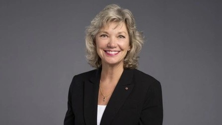 Smiling Kim Orth with short blond hair wearing a black blazer over a white top against a gray background.