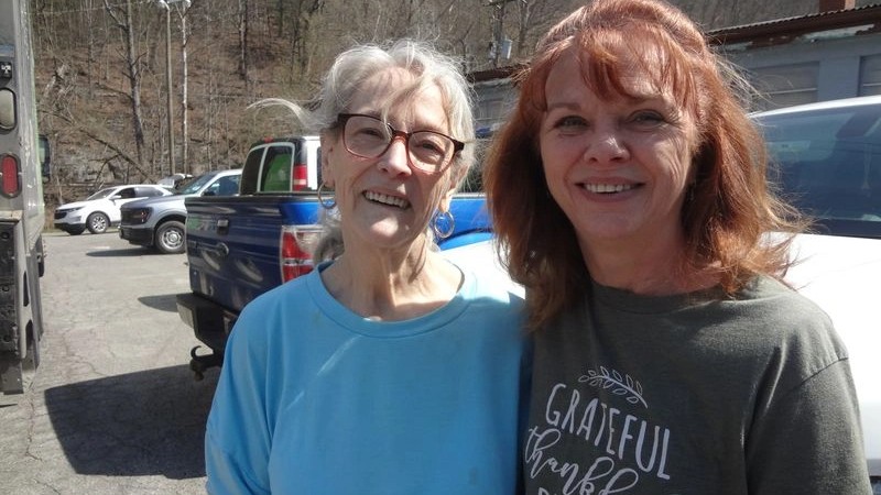 Two women smiling outdoors in a sunny parking lot, surrounded by cars.