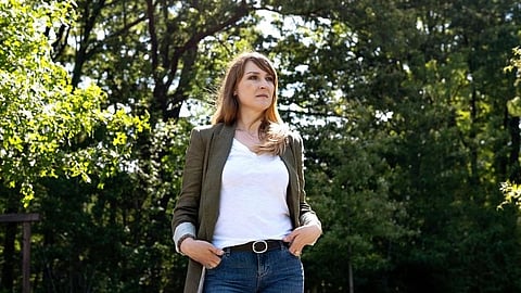 A woman stands confidently outdoors, wearing a white shirt and olive jacket, hands in pockets.
