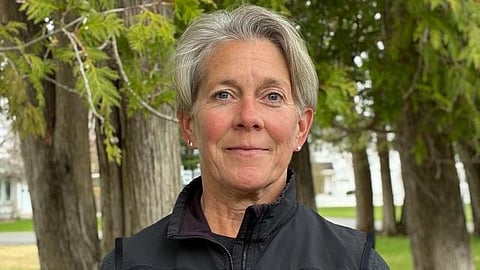 A person with short gray hair wearing a black vest stands in front of trees.
