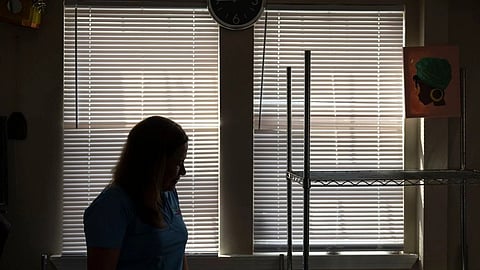 A woman in a blue shirt stands by a window, looking outside with a thoughtful expression.