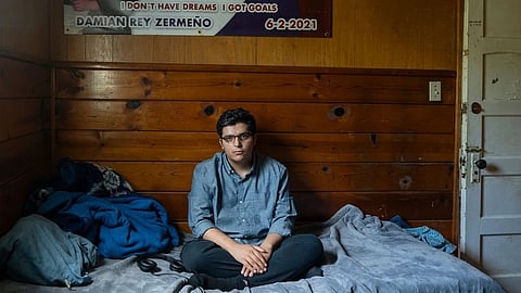 A man sits on a bed in a room, with a colorful poster hanging on the wall behind him.
