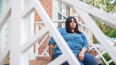A woman sits quietly on her front porch steps, wearing a blue shirt and jeans.