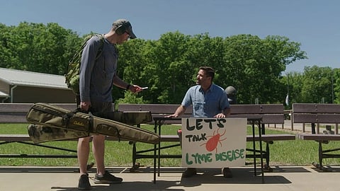 A man talks to another man holding a “Let’s Talk Lyme Disease” sign outdoors on a sunny day.