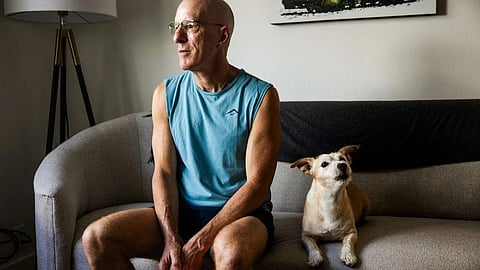 A bald man with glasses sits on a sofa beside a small dog.
