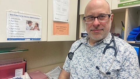 A pediatrician stands in a clinic with vaccine information displayed in the background.