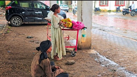 A man sitting next to the corpse of his wife on a stretcher and their daughter standing in front.