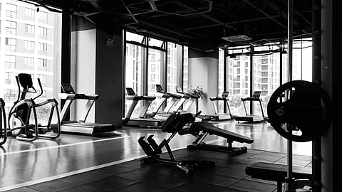 Black and white image of an empty gym with row of treadmills by large windows.
