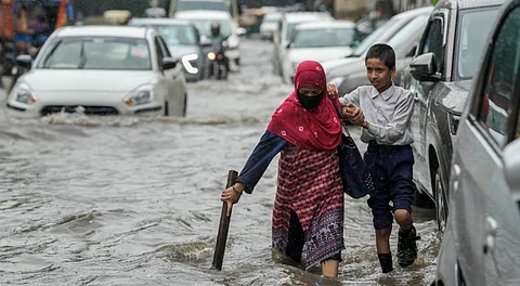 A woman, accompanied by her school-going child, uses a wooden stick to wade through a waterlogged road amid rainfall, in New Delhi, Thursday, Aug. 14, 2025.