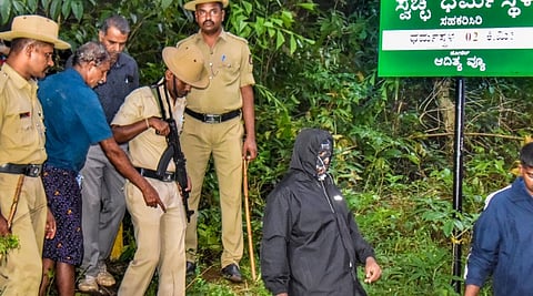 Security personnel with the whistleblower, in mask, near a site of an alleged burial related to the Dharmasthala mass burial case, at Dharmasthala, in Dakshina Kannada district.