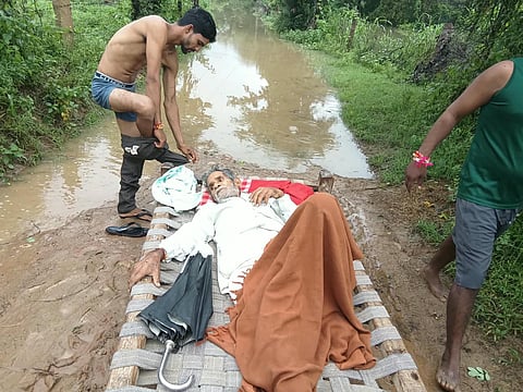 Villagers Carry a Sick Elderly Man on a Cot Across a Flooded Culvert