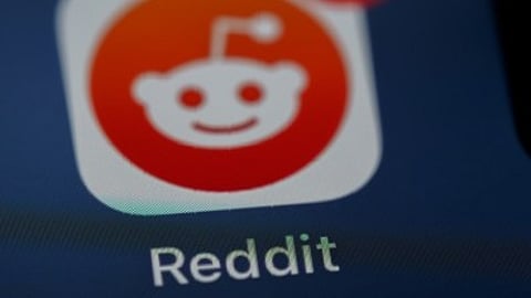 Close-up of the Reddit app icon on a smartphone screen, featuring the iconic alien logo in white on an orange circle, with "Reddit" below.