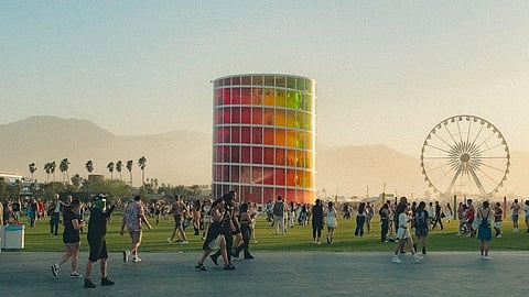 A colorful cylindrical art structure at a festival, with people walking around and a Ferris wheel in the background.