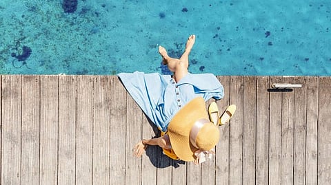 A person sitting on a wooden deck with their feet in a blue pool, wearing a hat and a light dress.