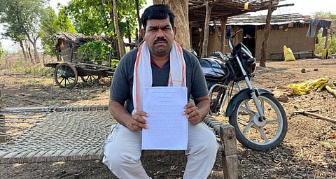 Man sitting on a cot in a rural setting holding a paper, with a motorcycle and huts in the background