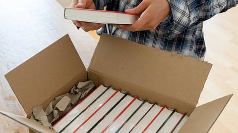 Person holding a book beside an open box filled with stacked books.”