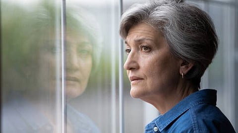 An elderly woman looking out of a window with her reflection visible on the glass.