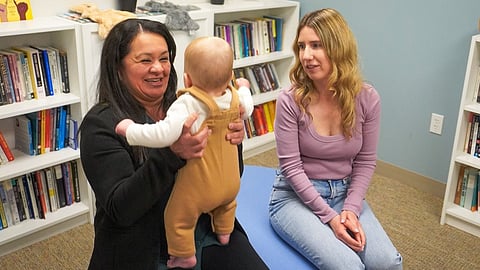A woman cradles a baby while another woman holds a book, both engaged in a moment of connection and learning