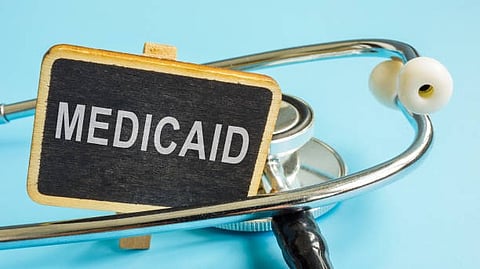 A small chalkboard sign with the word "Medicaid" is placed in front of a stethoscope on a light blue background.