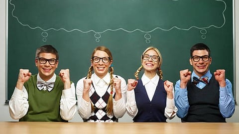 Four smiling students in glasses posing enthusiastically in front of a chalkboard.
