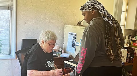 Two people talking at a desk, one older person sitting and the other standing beside them.