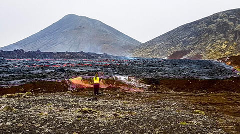 Person standing near flowing lava with a volcano in the background.