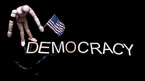 A wooden mannequin holding a small U.S. flag above the word “DEMOCRACY.”