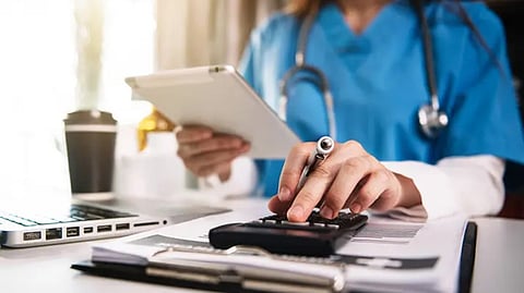 A doctor working on a laptop while holding a clipboard and pen.