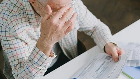 The image shows an elderly person in a checkered shirt holding their face while sitting at a table with documents.