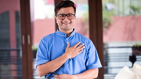 The image shows a smiling man in blue medical scrubs with a stethoscope, standing with one hand on his chest.
