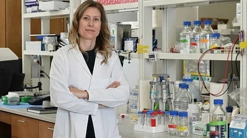 The image shows a woman in a white lab coat standing confidently in a laboratory filled with scientific equipment and bottles.