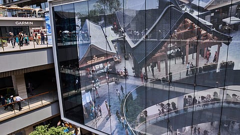 Modern shopping mall with glass architecture, outdoor walkways, and people walking around retail stores.