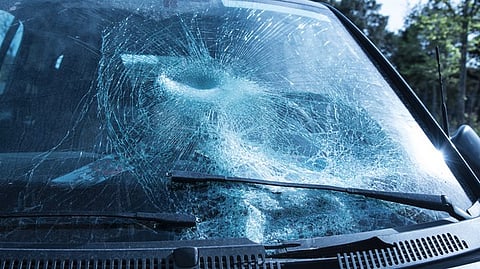 A close-up view of a car windshield badly cracked and shattered, showing a large impact mark near the center, likely from a strong collision or object hit.