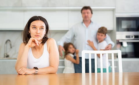 Woman sitting at a table looking thoughtful, with a man and two children in background.