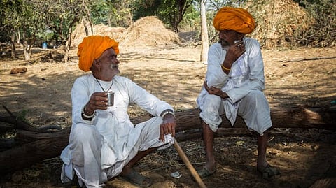 Two men in taditional attire with orange turbun siiting outdoors and talking.
