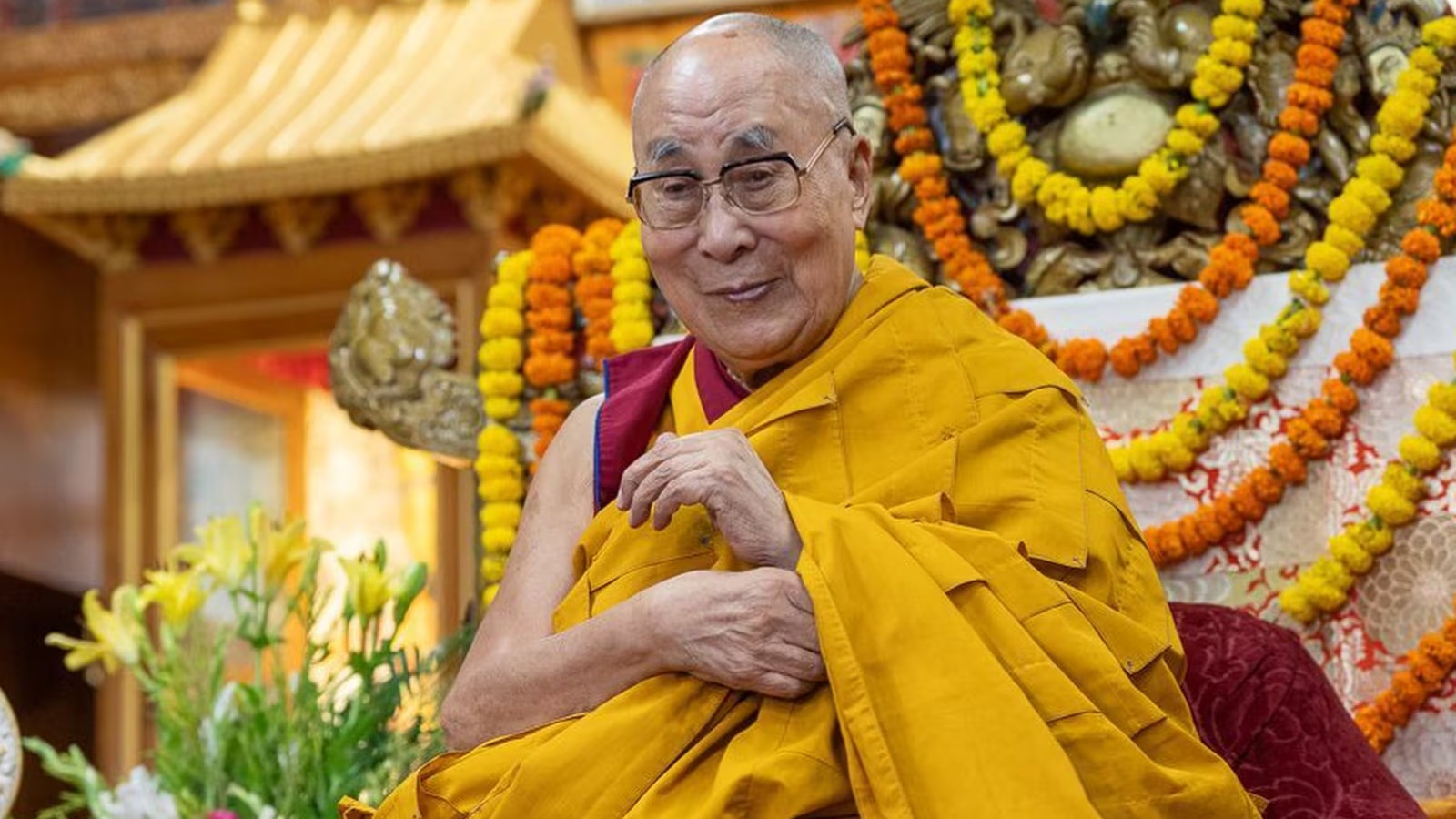 A budhhist monk wearing yellow and red robes is seated in front of a decorated background with marigold garlands and flowers.