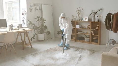 A person wearing a full white protective suit and blue shoe covers is cleaning or inspecting the floor in a modern, minimalistic room with wooden furniture and indoor plants