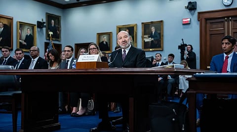 An elderly man speaks at a podium in a formal meeting room filled with seated attendees, with framed portraits hanging on the walls behind him.