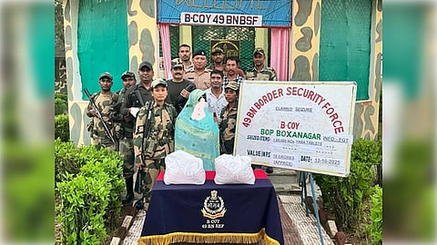 A group of Border Security Force personnel stands in front of a camouflage-painted building with a "49 BN BSF" sign. They pose with seized goods and a sign detailing their location and operation. The atmosphere is serious and focused.