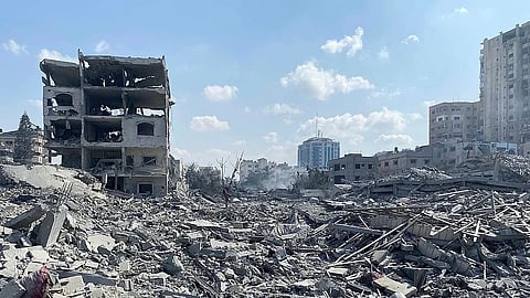 Rubble and debris cover the ground of a city, with partially destroyed buildings in the background under a bright blue sky with scattered clouds.