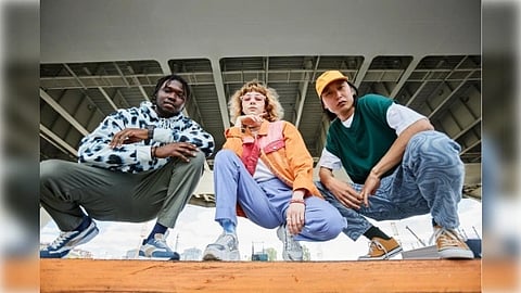 Three individuals in vibrant streetwear crouch confidently under an urban bridge. The setting is dynamic, conveying a sense of style and camaraderie.