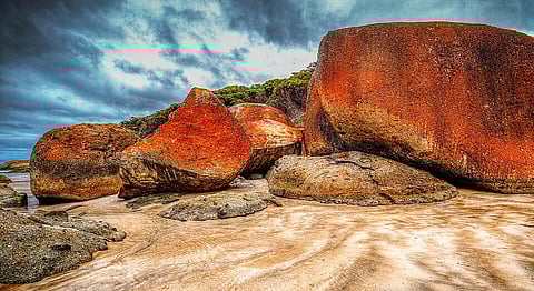 Dramatic coastal scene with large, vibrant orange and grey boulders on a sandy beach. A moody, overcast sky enhances the rugged, natural beauty.