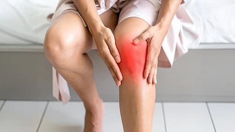 A person sitting on a bed holds their knee, which is highlighted in red, indicating pain or inflammation. The setting is indoors, with a neutral tone.