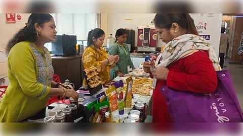 A woman in a red outfit examines products at a market stall staffed by women in colorful attire. The table is filled with assorted packaged goods.