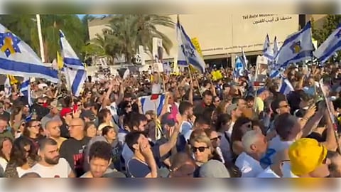 A large crowd protests outside the Tel Aviv Museum, waving Israeli flags. The atmosphere is energetic and peaceful, under a clear sky.