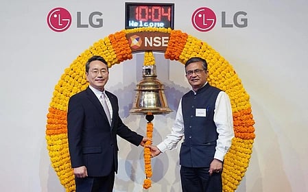 Two men stand smiling, ringing a large ceremonial bell adorned with yellow and orange flowers. LG logos are visible above their heads, symbolizing a celebratory occasion.