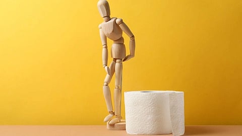 A wooden mannequin leans forward, holding its stomach, next to a roll of toilet paper. The background is a plain yellow, conveying a humorous tone.